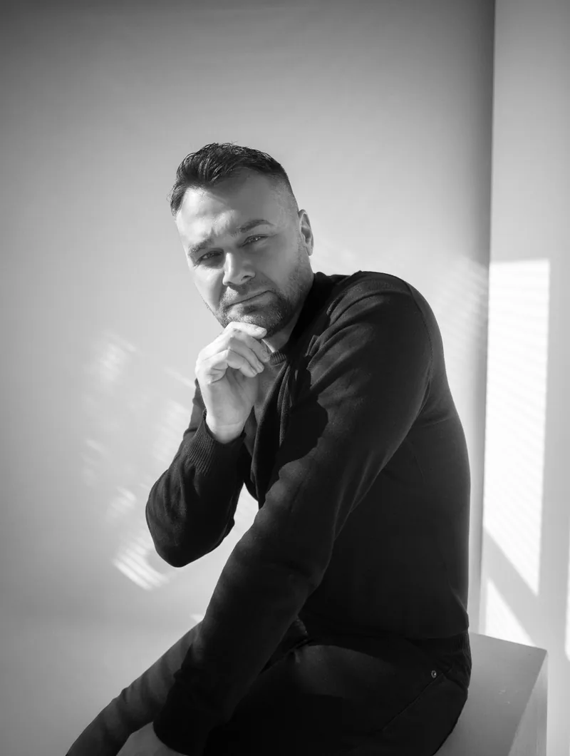 Pavel Filandr v černém svetru sedí v denním světle u okna, černobílý portrét